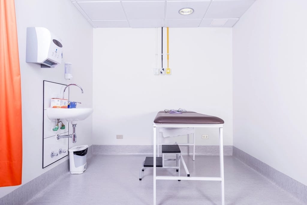 Une salle d'examen médical vide, avec une table d'examen, un marchepied et un lavabo, sous une lumière blanche.